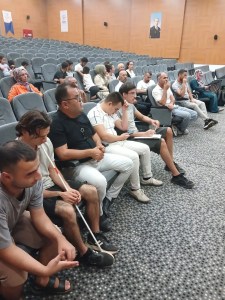 Fotoğrafta bir konferans salonunda oturan bir grup insan görülüyor. Salonun koltukları gri renkte ve oldukça fazla sayıda boş koltuk var. Ön sırada oturan kişiler arasında bir beyaz baston tutan bir adam dikkat çekiyor. Çeşitli yaş gruplarından insanlar var ve çoğu kişi dikkatle bir şeye odaklanmış gibi görünüyor. Arka planda, duvarda Atatürk'ün bir portresi asılı.