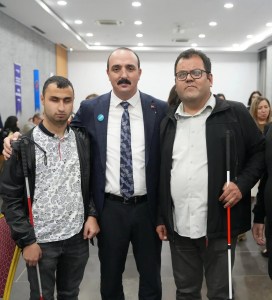 Üç kişi bir arada poz veriyor. Ortadaki kişi Konyaaltı belediye başkanı Cem KOTAN takım elbise giymiş, kravat takmış ve yakasında mavi bir rozet var. Sağındaki ve solundaki Kişiler beyaz baston tutuyorlar. Sol taraftaki kişi Mert YAKIŞTIR desenli bir tişört ve siyah bir ceket giymiş. Sağ taraftaki kişi ise Oğuz ÖZDEMİR beyaz bir gömlek ve siyah bir ceket giymiş. Arka planda başka insanlar da var ve ortam kapalı bir alan gibi görünüyor. Duvarlarda asılı afişler var.